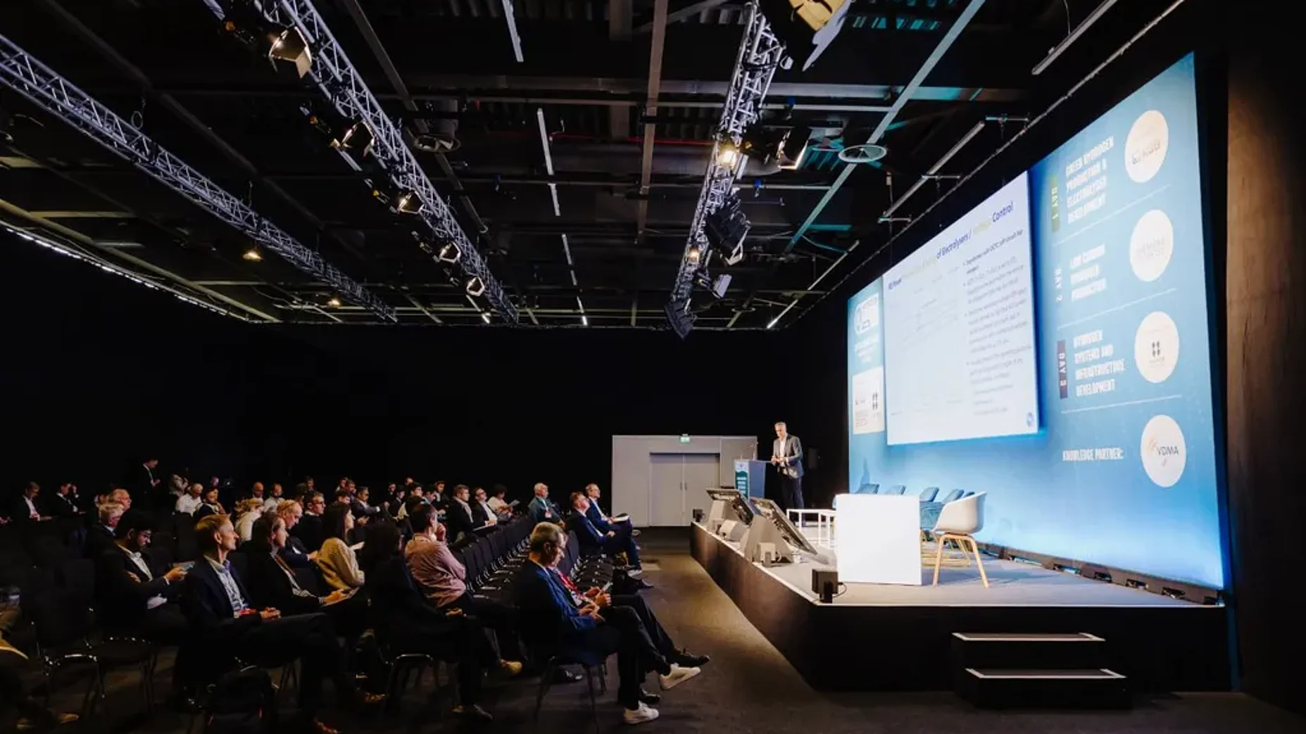 Eine Person hält auf einer Konferenzbühne einen Vortrag vor einem sitzenden Publikum.